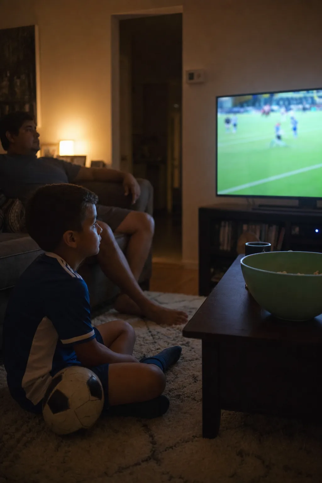 A kid in a soccer jersey sits on the living room rug watching a World Cup match, a ball at their knee